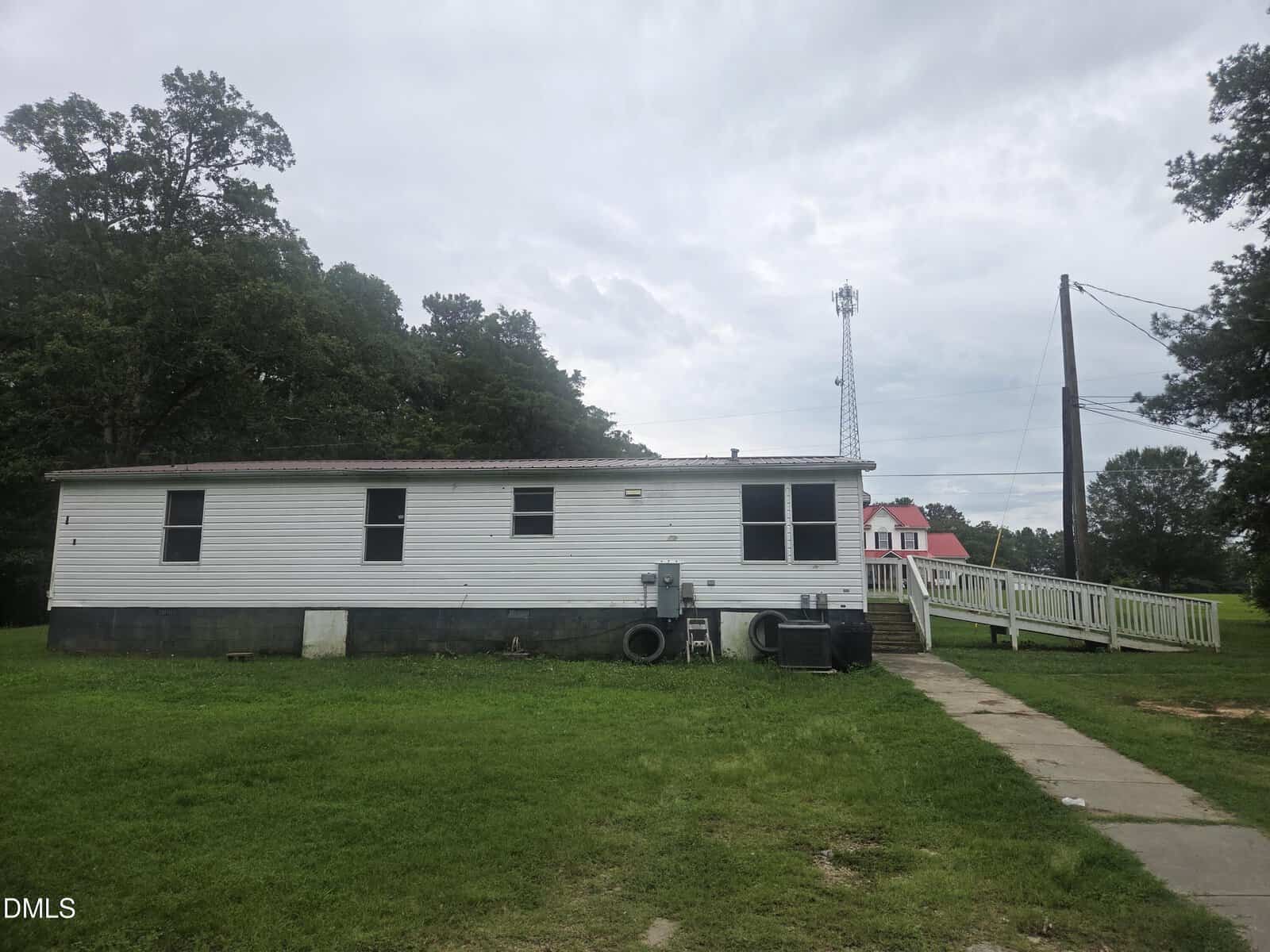 A white mobile home with a wheelchair ramp sits on a grassy yard. Trees are in the background, and a red-roofed house and cell tower are visible in the distance under a cloudy sky.