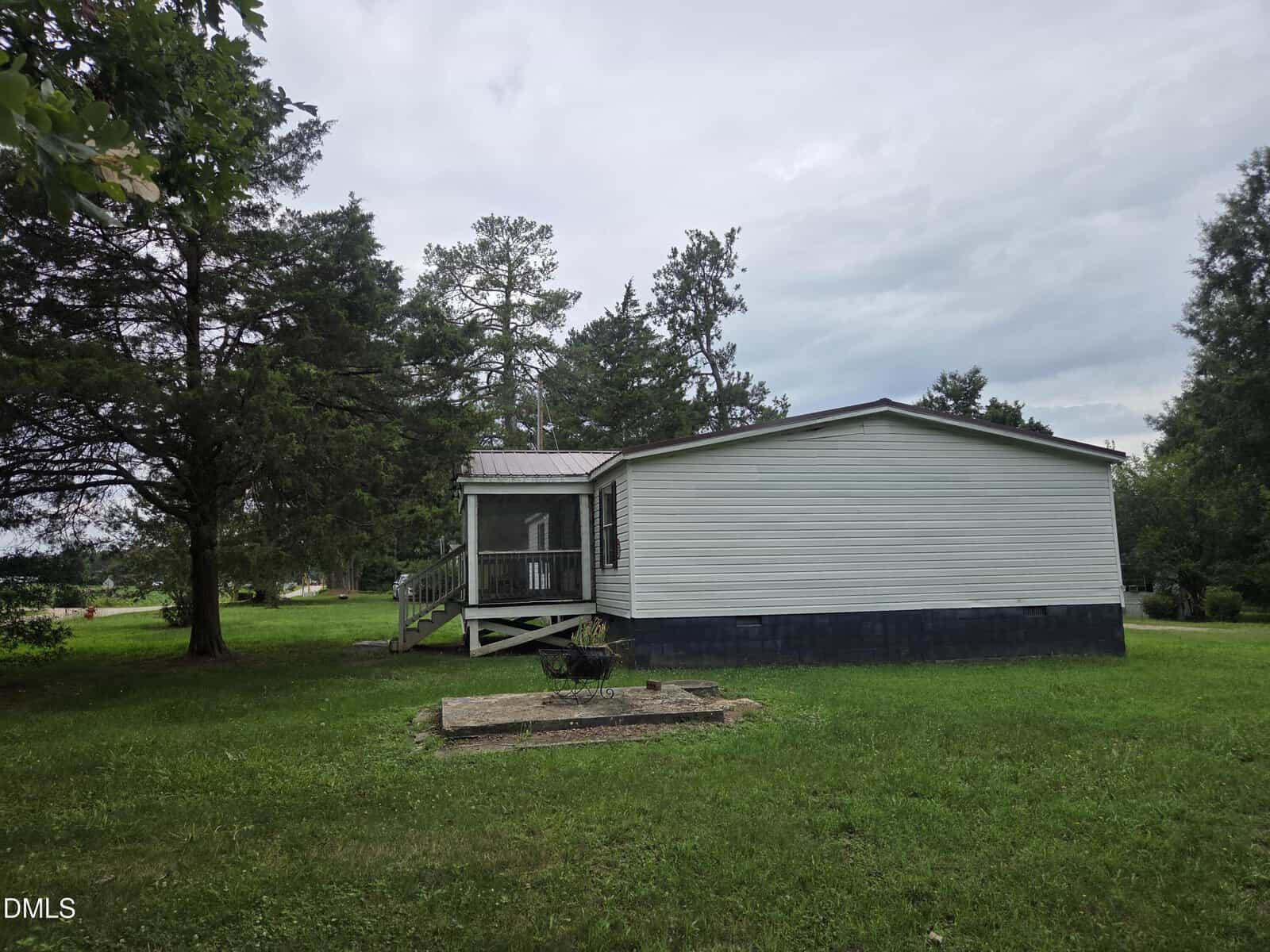 A white manufactured home with a screened porch sits on a green lawn, surrounded by trees under a cloudy sky. A small concrete pad is in the grass near the home.