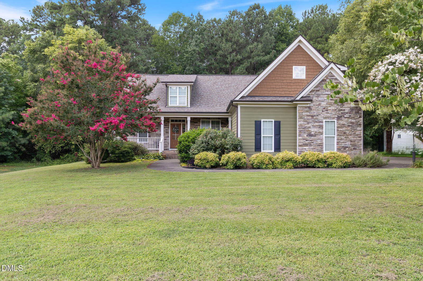 Charming single-story house with stone and green siding located at 2117 Olde Brassfield Drive, Franklinton. Featuring a welcoming front porch, gabled roof, blooming trees, and a large manicured lawn, this home is surrounded by tall trees and lush greenery in a quiet suburban setting.