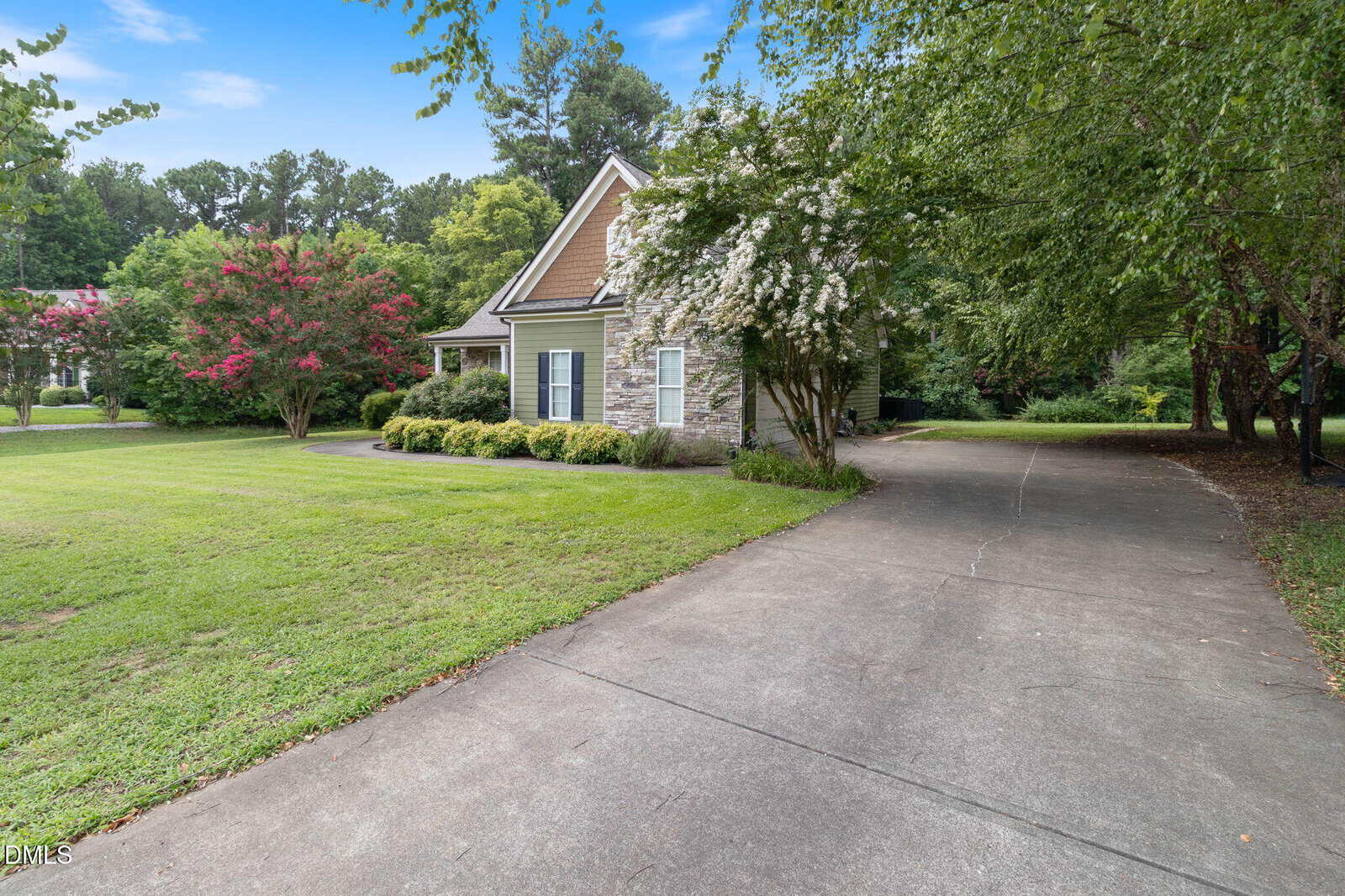 A stone and siding house with a gabled roof sits beside a long driveway, surrounded by green grass, flowering trees, and tall woods under a blue sky.
