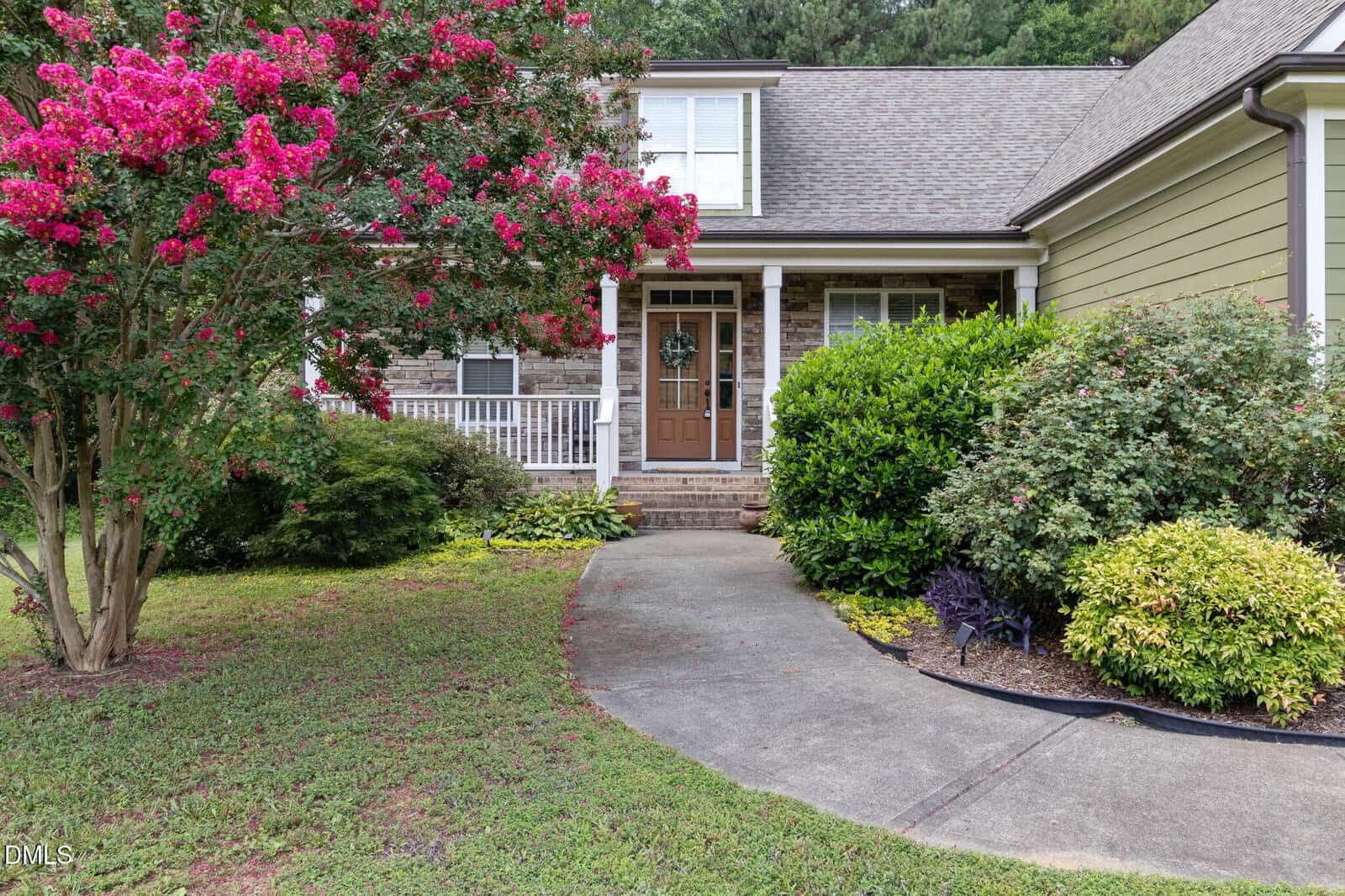 Curved concrete walkway leads to a house with a brick porch, white railing, green siding, and lush landscaping featuring shrubs and a flowering tree with bright pink blossoms.