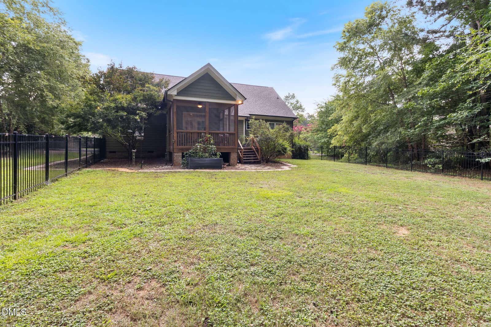 A backyard with green grass, a black metal fence, and a house featuring a screened porch. Trees and shrubs border the yard under a blue sky.