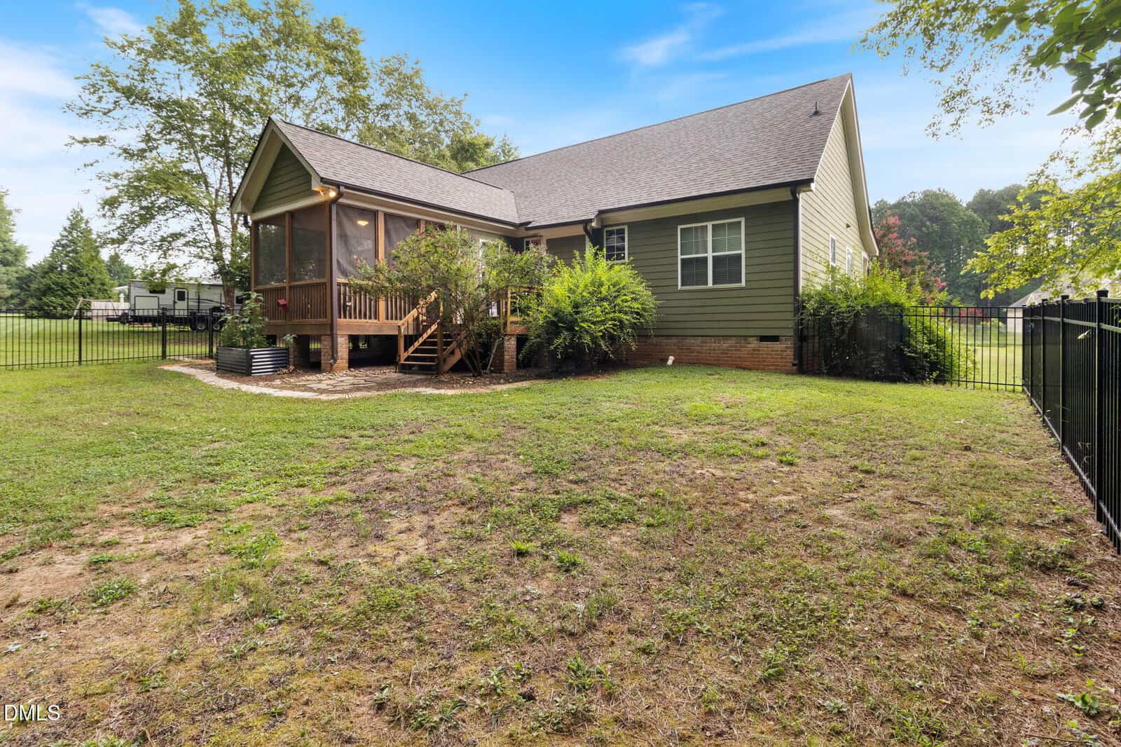 A green house with a screened porch, wooden steps, and a fenced backyard featuring grass, shrubs, and trees under a blue sky.