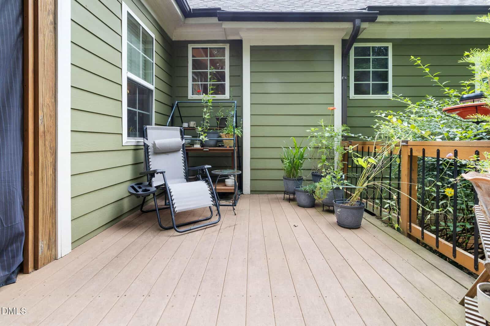 A small wooden deck with a reclining chair, potted plants, a shelving unit, and green siding on the house. Two windows and a railing are visible, creating a cozy outdoor space.