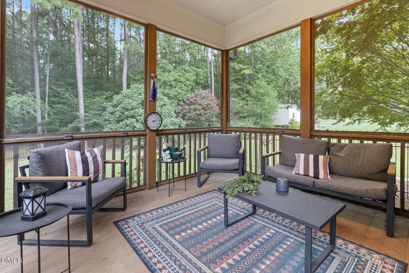 A cozy screened porch with gray seating, striped pillows, a patterned rug, and a black coffee table, surrounded by lush green trees and a grassy yard.
