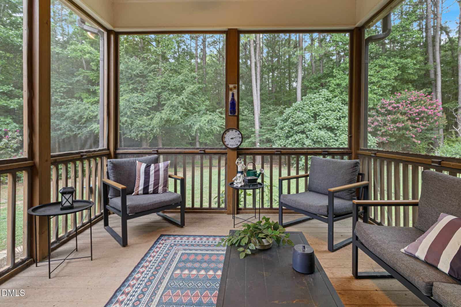 Screened-in porch with modern seating, patterned rug, coffee table with plants, clock on the wall, and a view of a lush green yard with trees and flowering bushes outside.