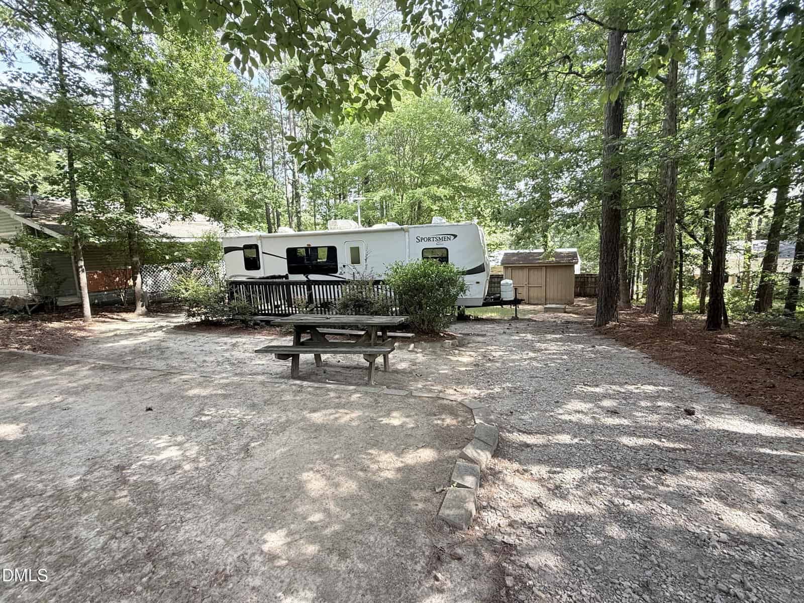 A white travel trailer is parked on a gravel lot surrounded by trees, with a wooden picnic table in the foreground and a small shed nearby. Shadows from the trees cover most of the area.