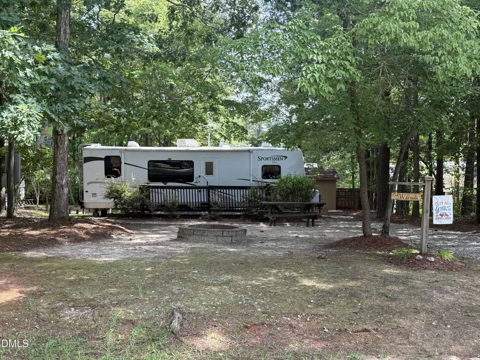 A white Sportsmen RV is parked in a wooded campsite at 126 Geronimo Drive, Louisburg, featuring a wooden deck, picnic table, small fire pit, and signs. Trees surround the area, providing shade and greenery.