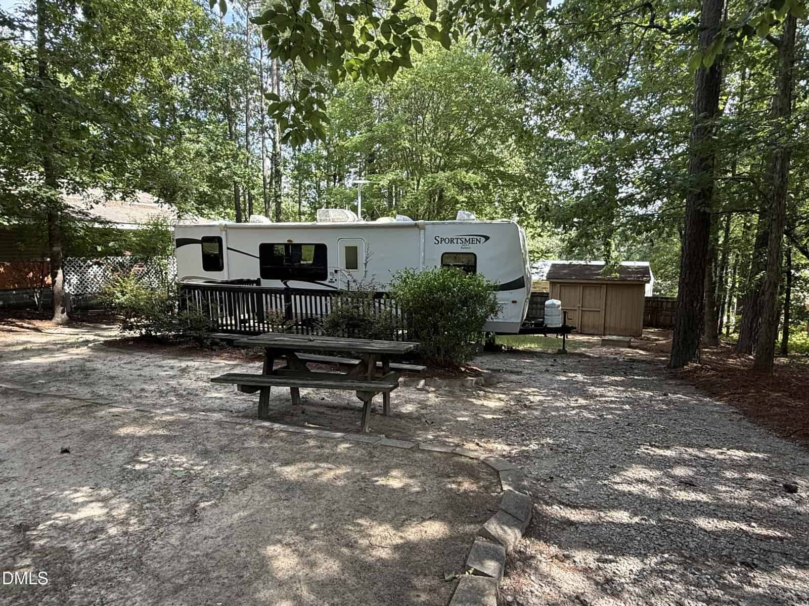 A white Sportsmen RV is parked among trees beside a small wooden shed. A picnic table sits on a gravel area in the foreground, surrounded by shaded woods.