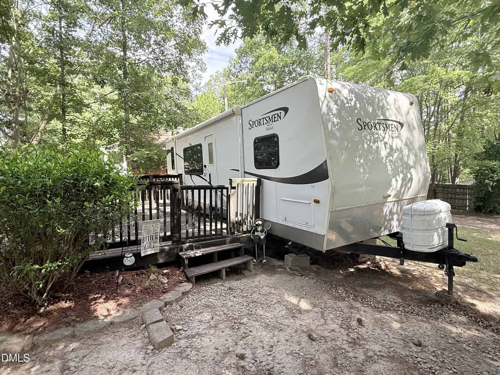 A white Sportsmen travel trailer is parked next to a wooden deck with steps, surrounded by trees and greenery in a shady outdoor setting.