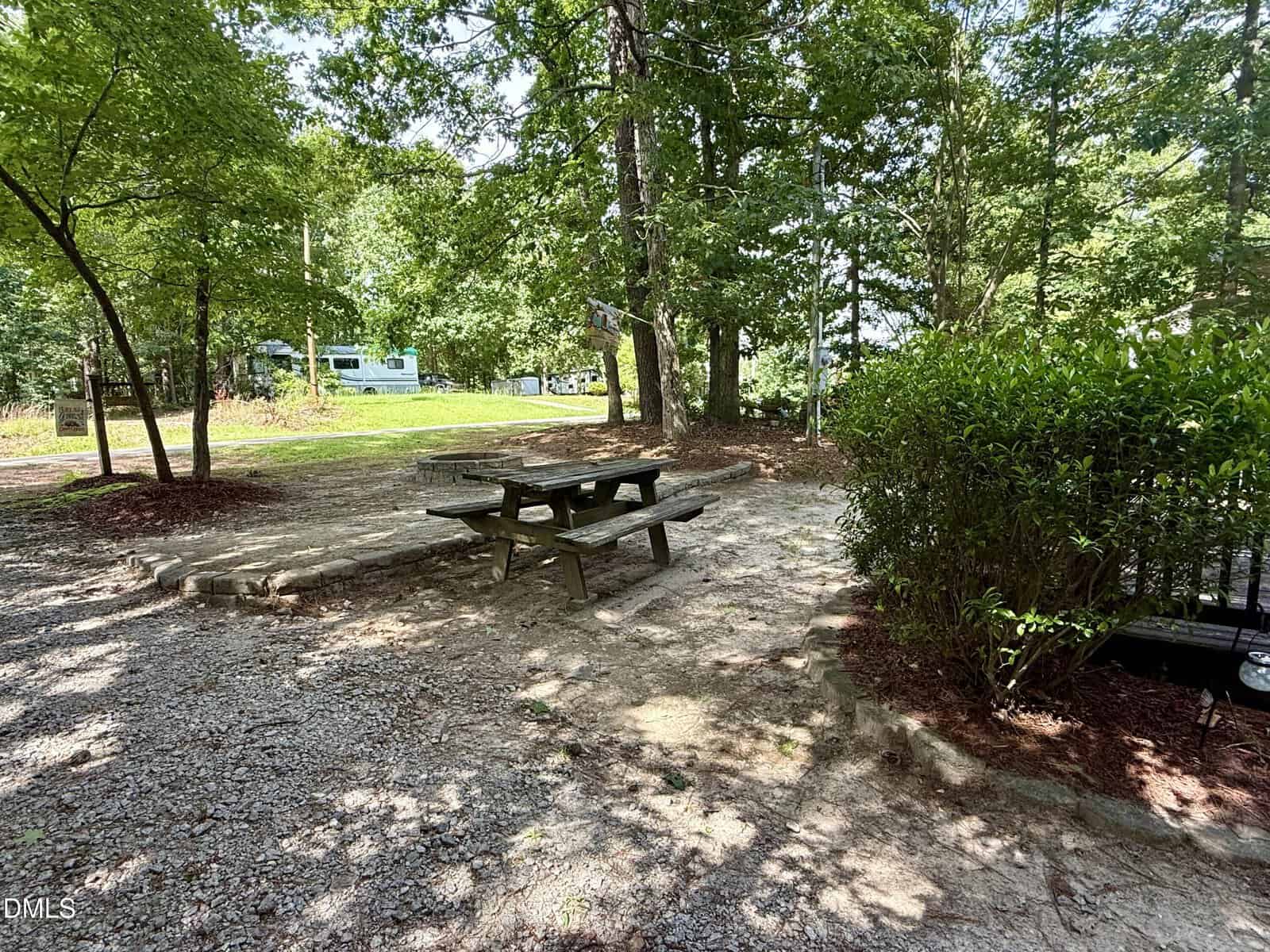 A wooden picnic table sits on a dirt and gravel area shaded by tall trees, with bushes nearby and a green lawn and house in the background.