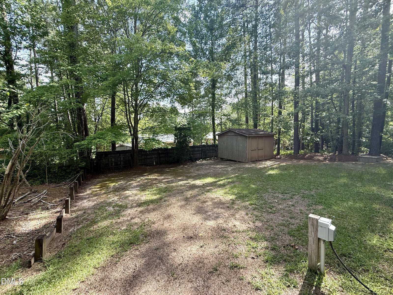 A fenced backyard with tall trees, a small wooden shed, a grassy area, and a gravel section. Sunlight filters through the trees, and a utility outlet is visible in the foreground.