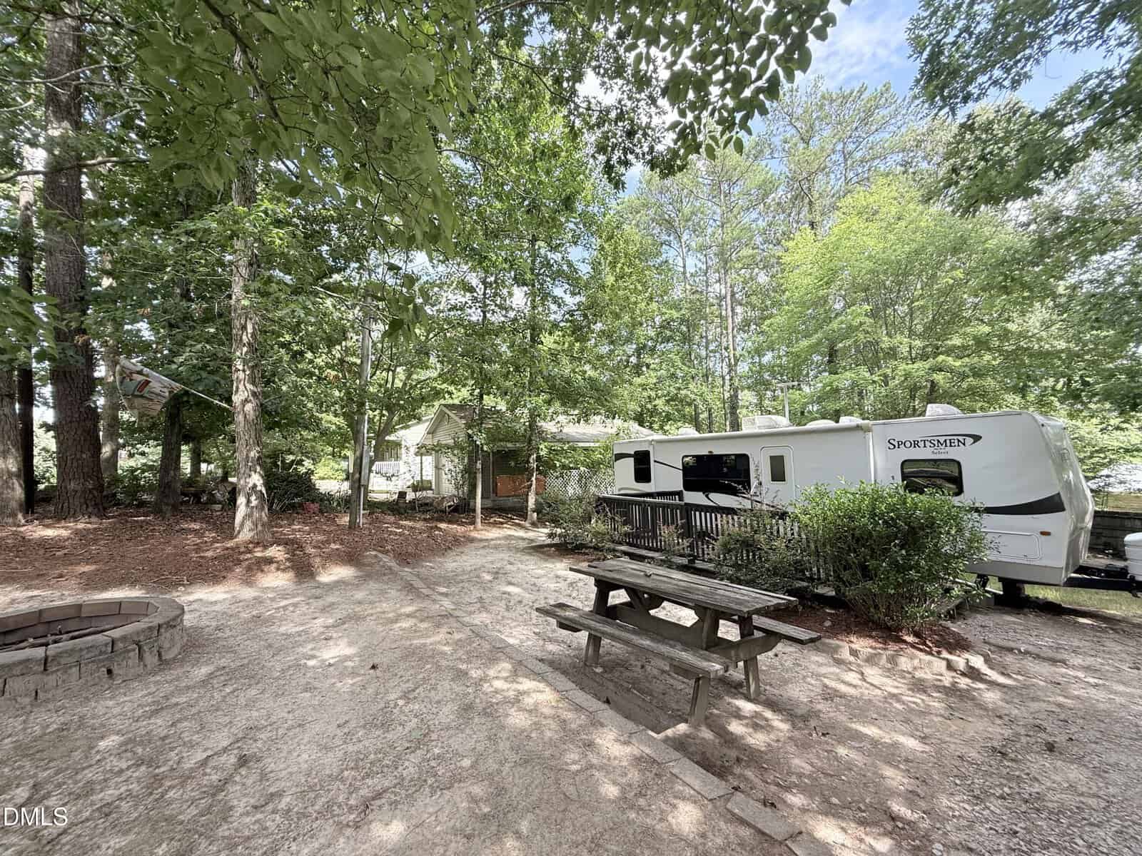 A white camper and a wooden picnic table sit in a shaded, tree-filled campsite. A fire pit is nearby, and a small house is visible in the background. Sunlight filters through the green leaves.