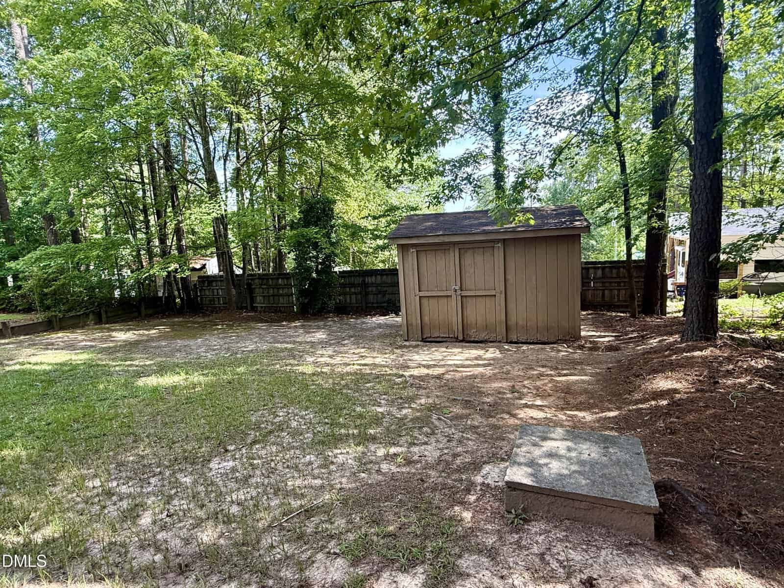 A wooden shed stands in a fenced backyard surrounded by tall trees. The ground is a mix of grass and bare patches, with a small concrete slab in the foreground. Sunlight filters through the trees.