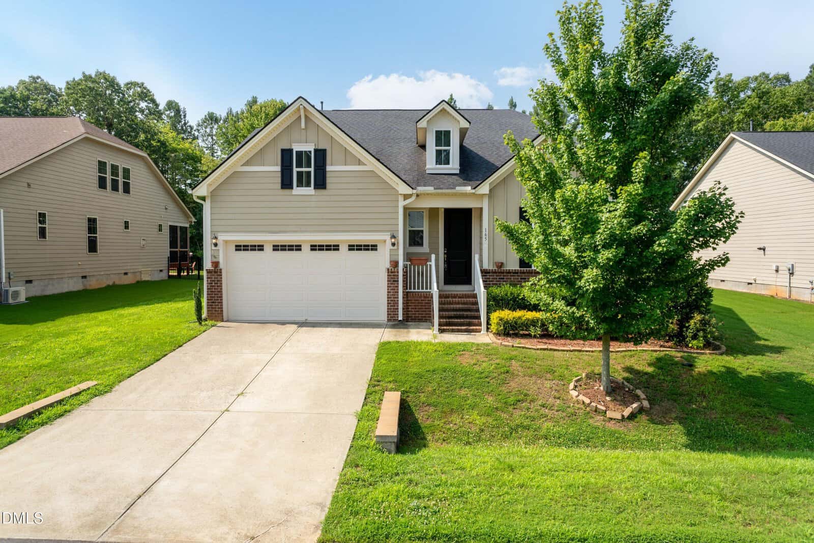 A beige single-family house at 165 Olde Liberty Drive in Youngsville features a two-car garage, brick accents, front porch, green lawn, small tree, and a concrete driveway, set between two similar houses on a sunny day.