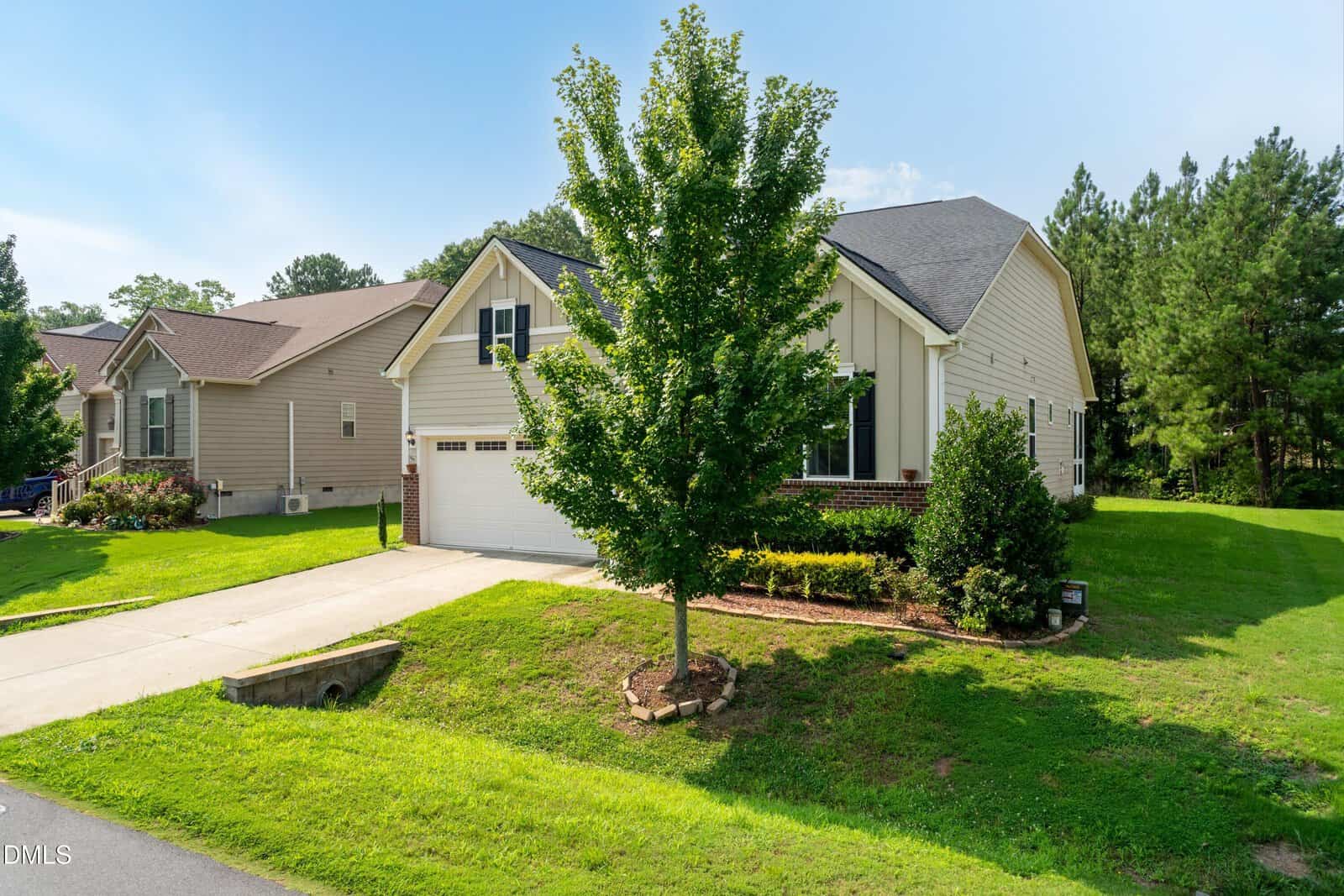 A beige single-story house with a two-car garage, a tree in the front yard, and a well-kept lawn, set in a suburban neighborhood with greenery and other homes nearby.