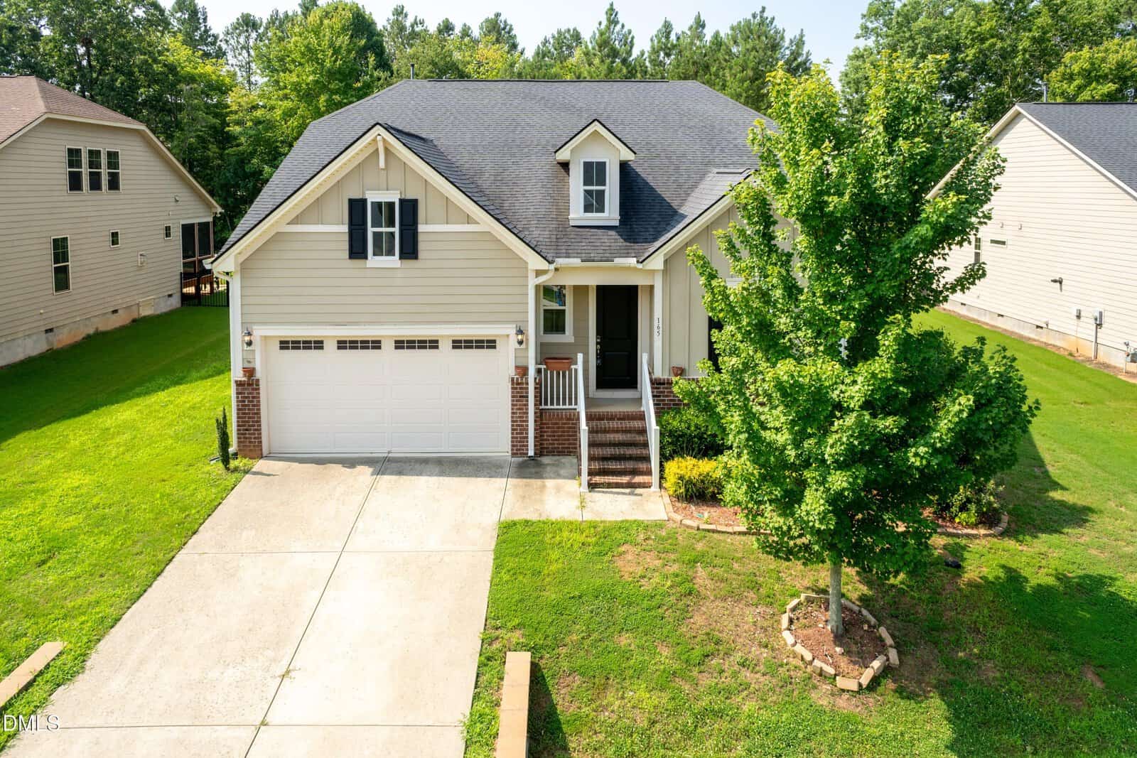 A beige single-family house with a dark gray roof, double garage, small front porch, brick accents, concrete driveway, and a well-maintained lawn with a young tree and neatly trimmed grass. Two neighboring homes are visible.