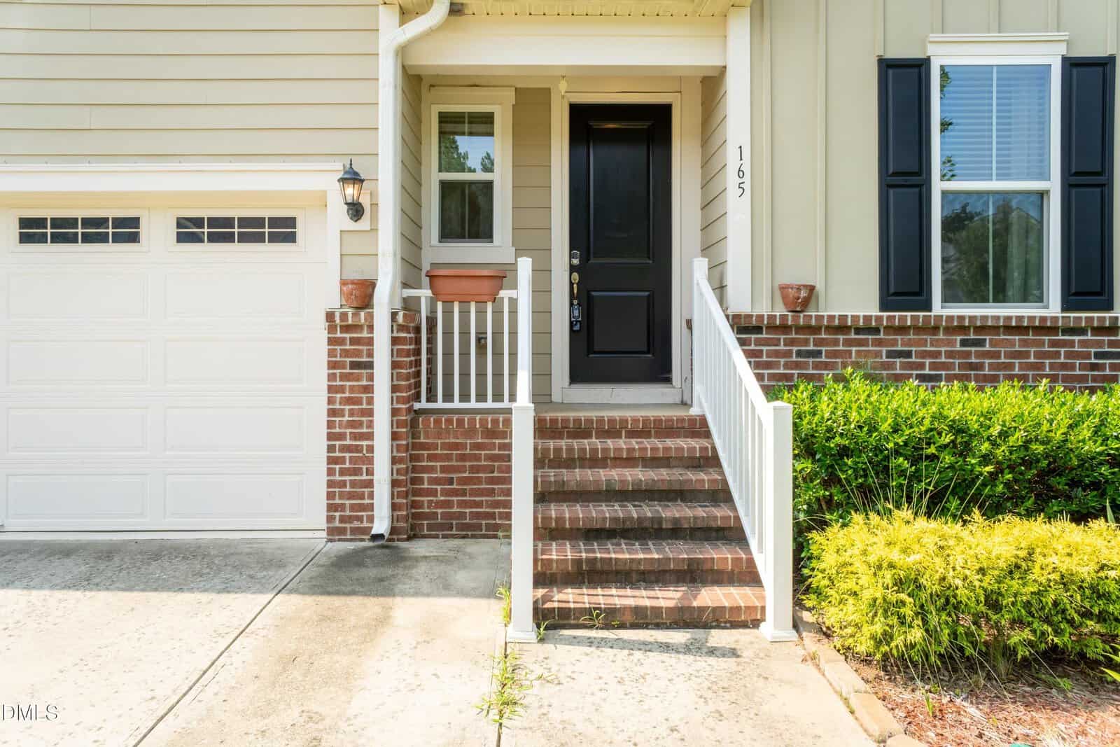Front entrance of a house with brick steps, white railing, black front door, beige siding, garage door on the left, and green bushes on the right.