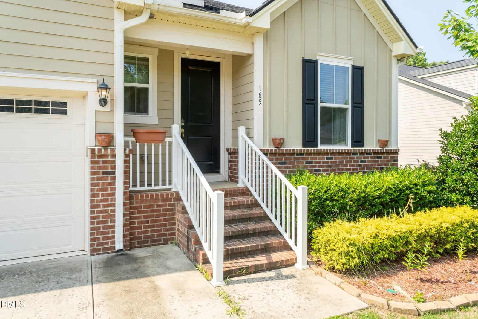 Front view of a house showing a small porch with steps, white railing, a black door, two windows with shutters, attached garage, brick and siding exterior, and landscaping with bushes and mulch.
