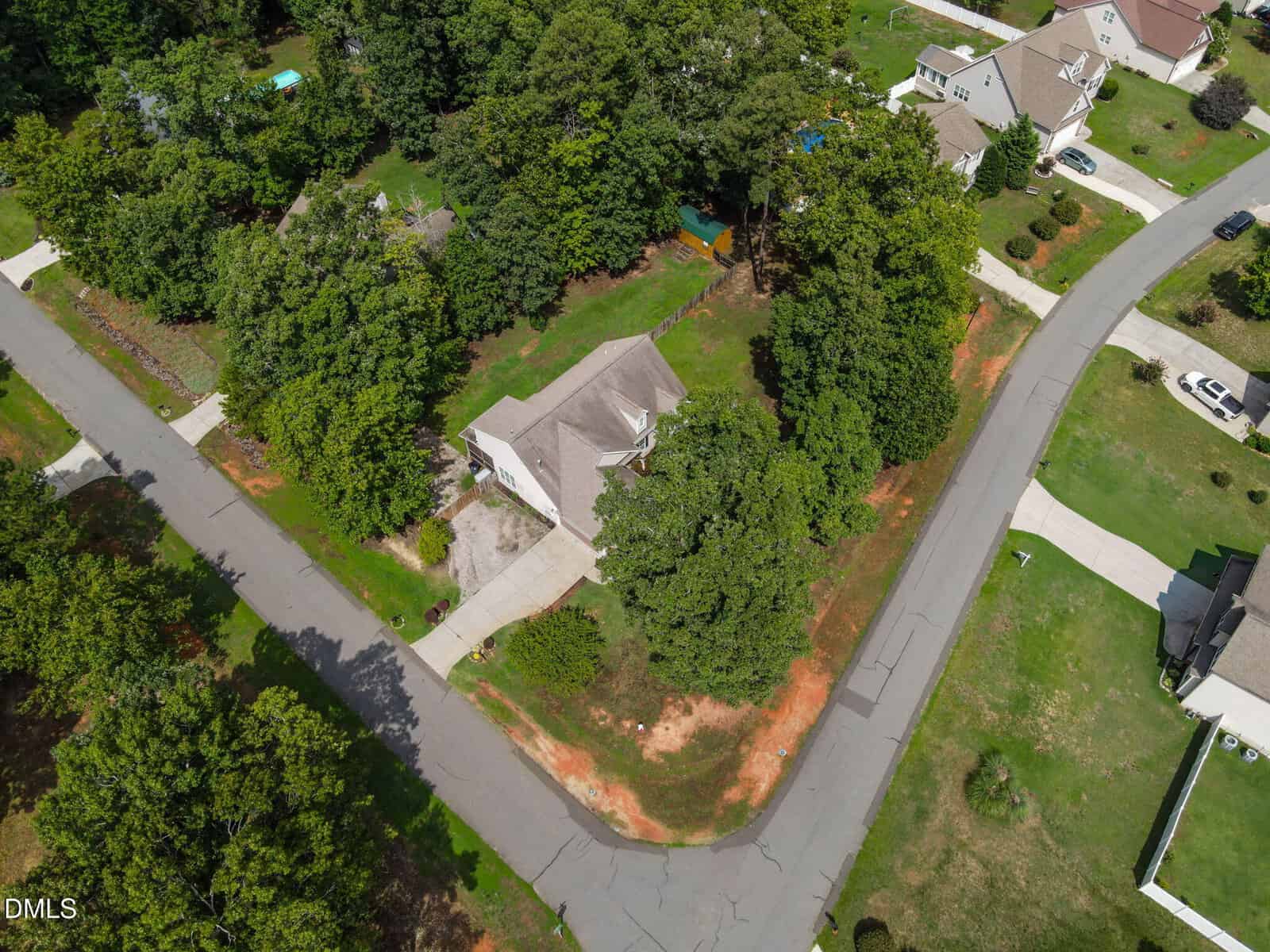 Aerial view of a house surrounded by trees at a street corner in a suburban neighborhood, with driveways, cars, green lawns, and nearby houses visible.