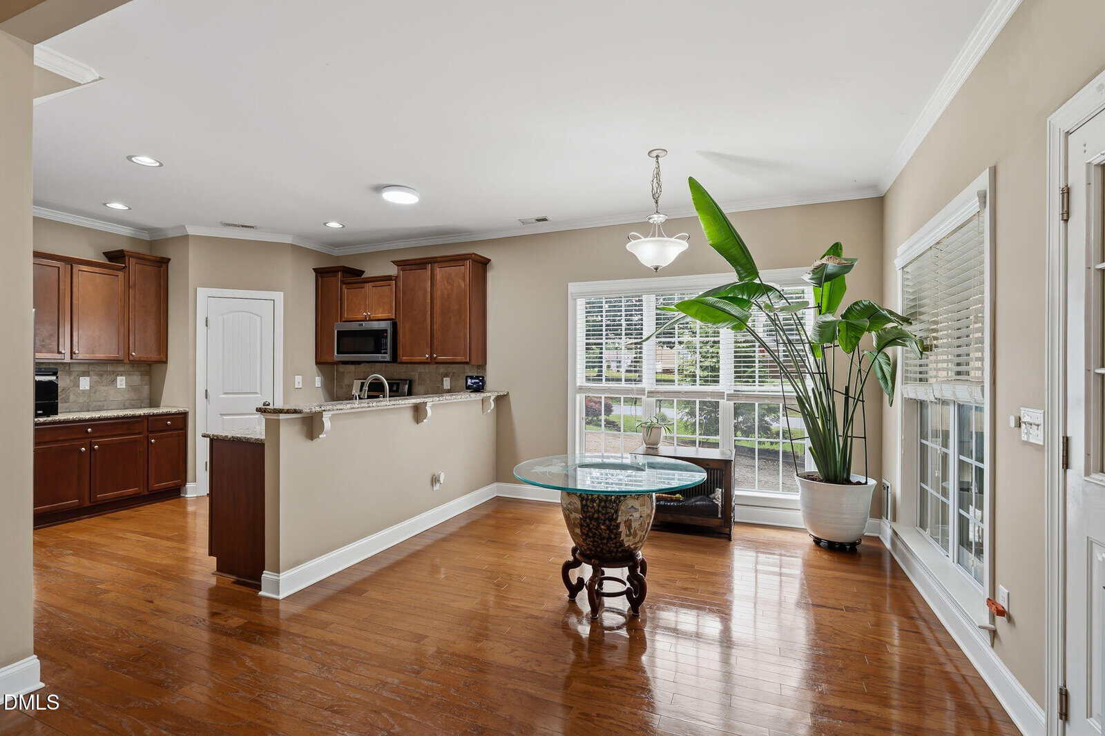 A modern kitchen with brown cabinets opens to a dining area with hardwood floors, a round glass table, large windows, a tropical plant in a pot, and neutral wall colors.