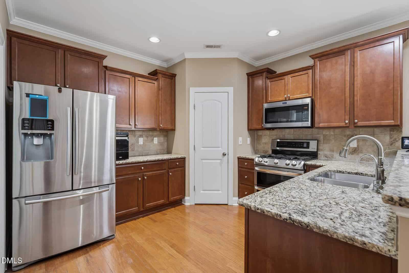 Modern kitchen with stainless steel appliances, granite countertops, brown wooden cabinets, a double sink, hardwood floors, and recessed lighting. White door in the center with beige walls and tile backsplash.