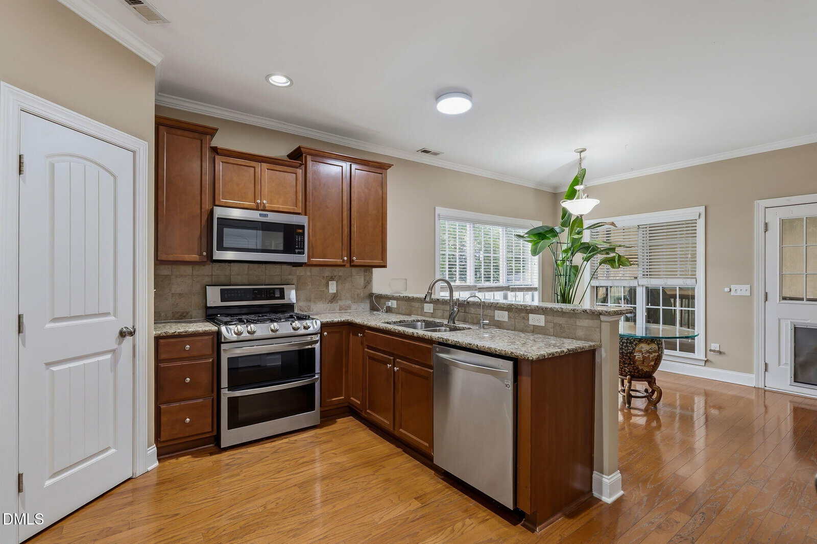 Modern kitchen with stainless steel appliances, brown cabinets, granite countertops, and wood flooring. There’s a large window, potted plant, and a bright open layout leading into another room with more windows.