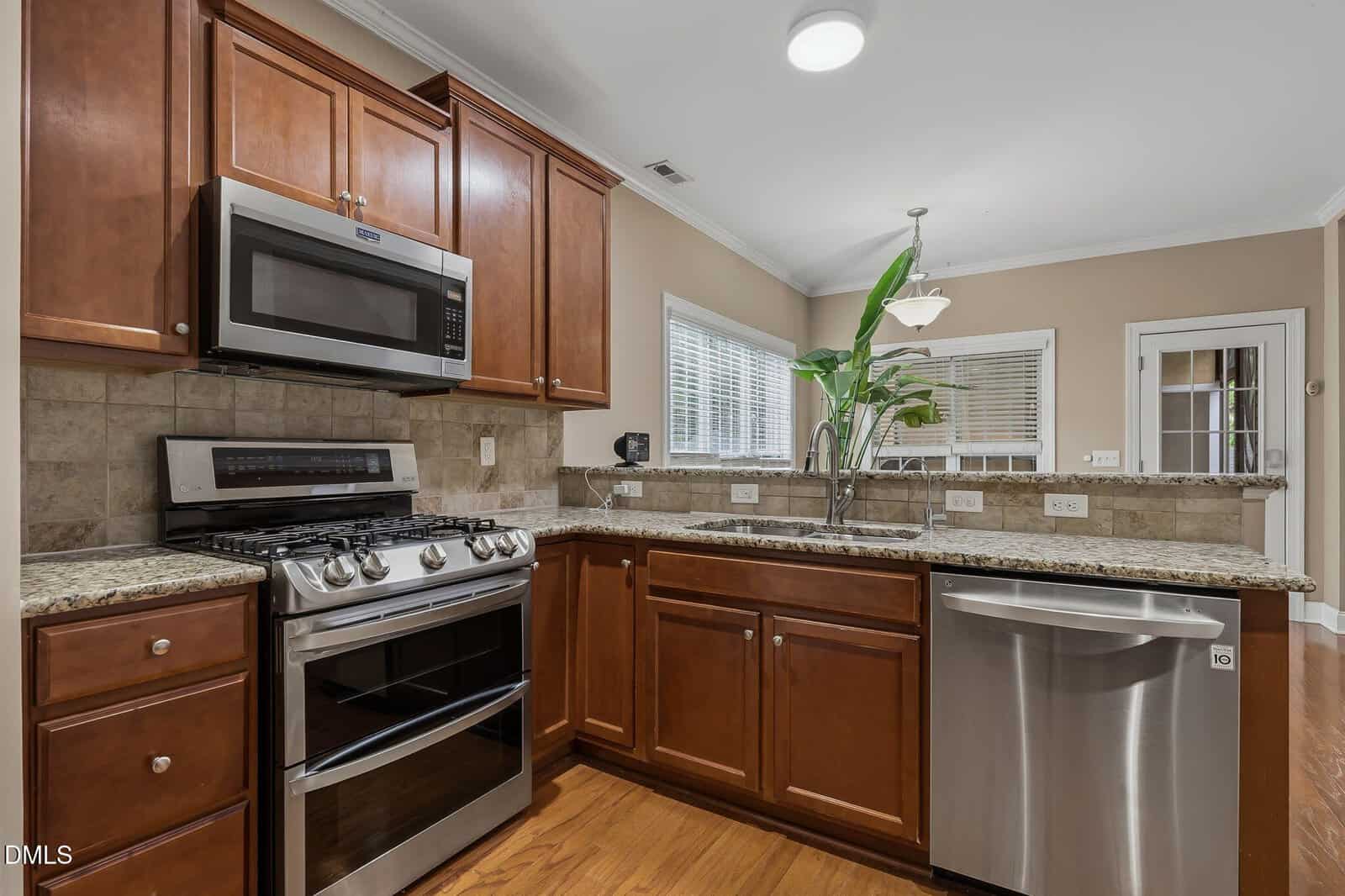 Modern kitchen with brown wooden cabinets, granite countertops, stainless steel appliances, gas stove, microwave, dishwasher, and a double sink. A large plant sits on the counter, and windows let in natural light.