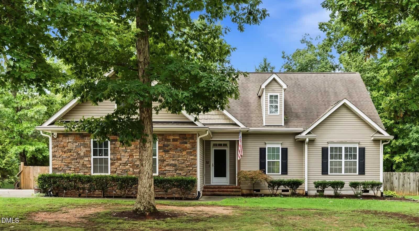 A beige house with stone accents, front porch, dormer window, and an American flag sits gracefully at 10 Madeira Drive in Franklinton. Green trees and shrubs frame the home, with a grassy yard in front.