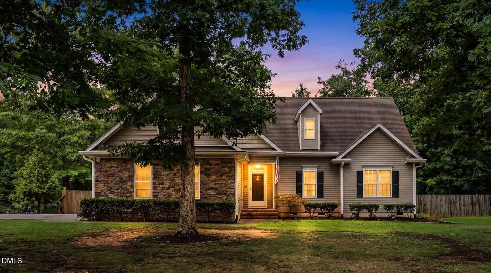 A two-story suburban house with lit windows and a stone facade sits behind a large tree and green lawn at dusk, with a glowing porch light welcoming visitors.
