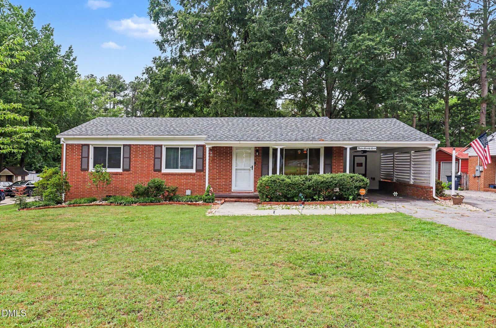 Charming single-story red brick house with a gray roof, white trim, and a carport on the right, located at 1277 David Avenue in Henderson. There are shrubs in front and a large front yard with a concrete path, surrounded by trees and greenery under a blue sky.