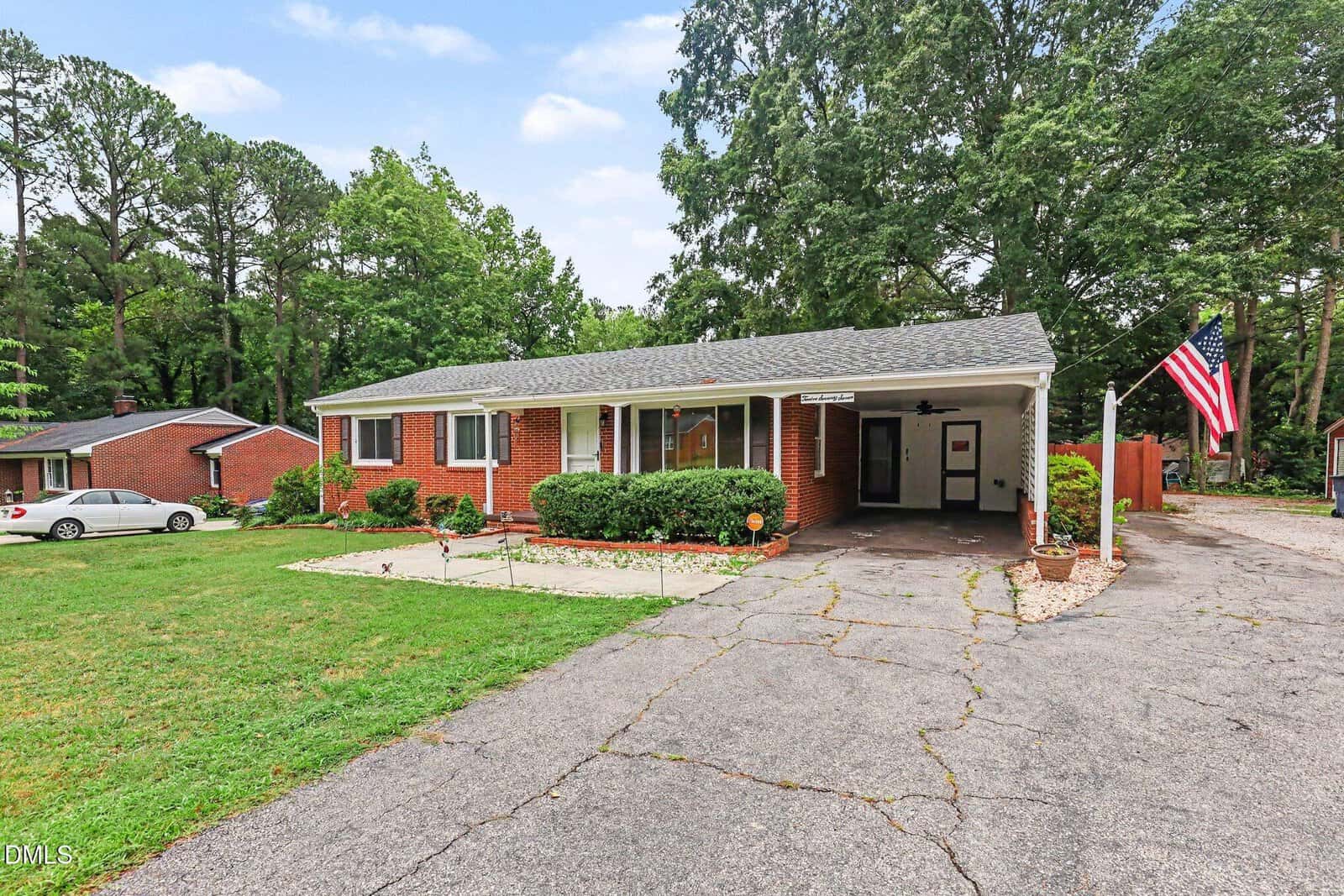 Single-story brick house with a carport, American flag, and manicured lawn, surrounded by trees and neighboring homes under a partly cloudy sky.