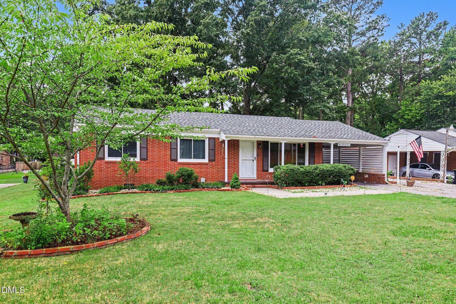 Single-story brick house with gray roof, white trim, and a covered carport. A green lawn with landscaped flower beds and trees is in front, with other houses and tall trees visible in the background.