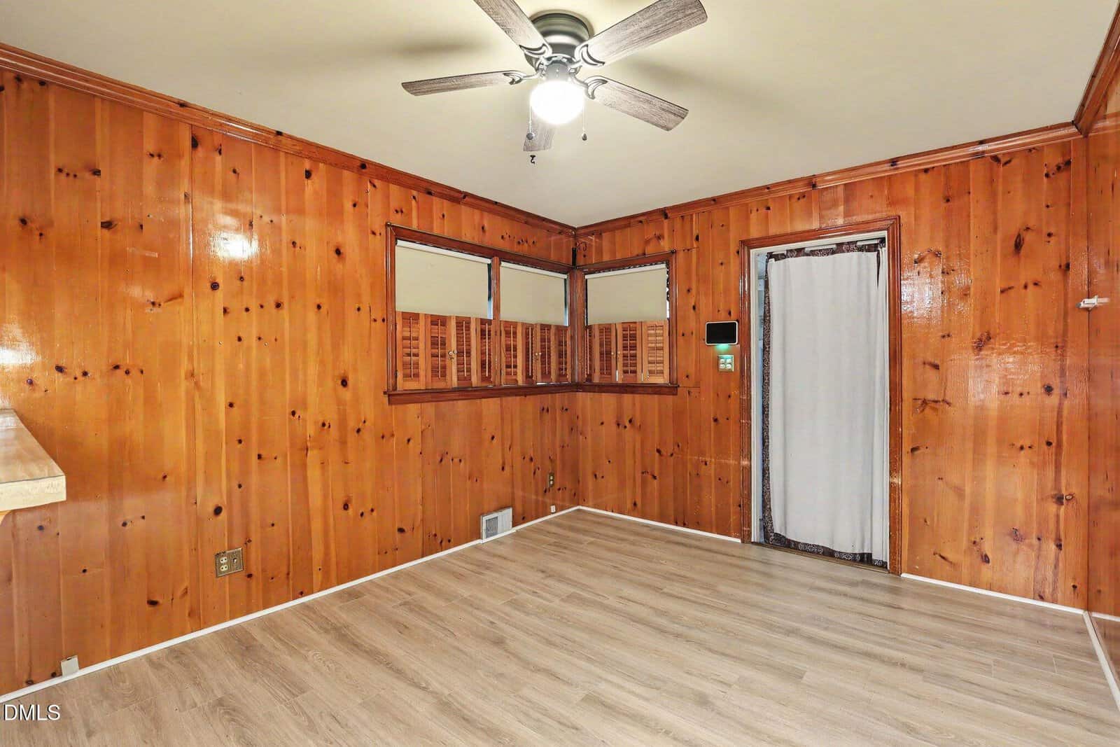 Room with wood-paneled walls, light wood flooring, windows with wooden shutters, a ceiling fan with lights, vent on the floor, and a door with a sheer curtain. Room is empty and well-lit.