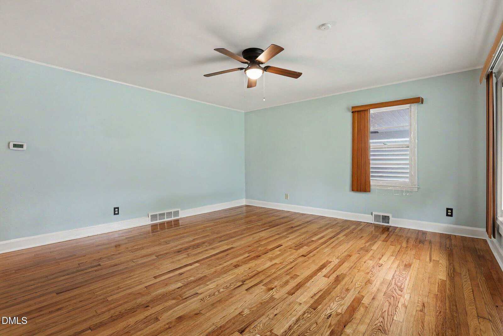 Empty room with light blue walls, a wooden floor, a ceiling fan with light, a window with a vertical blind, and two wall vents. No furniture or decor is present.