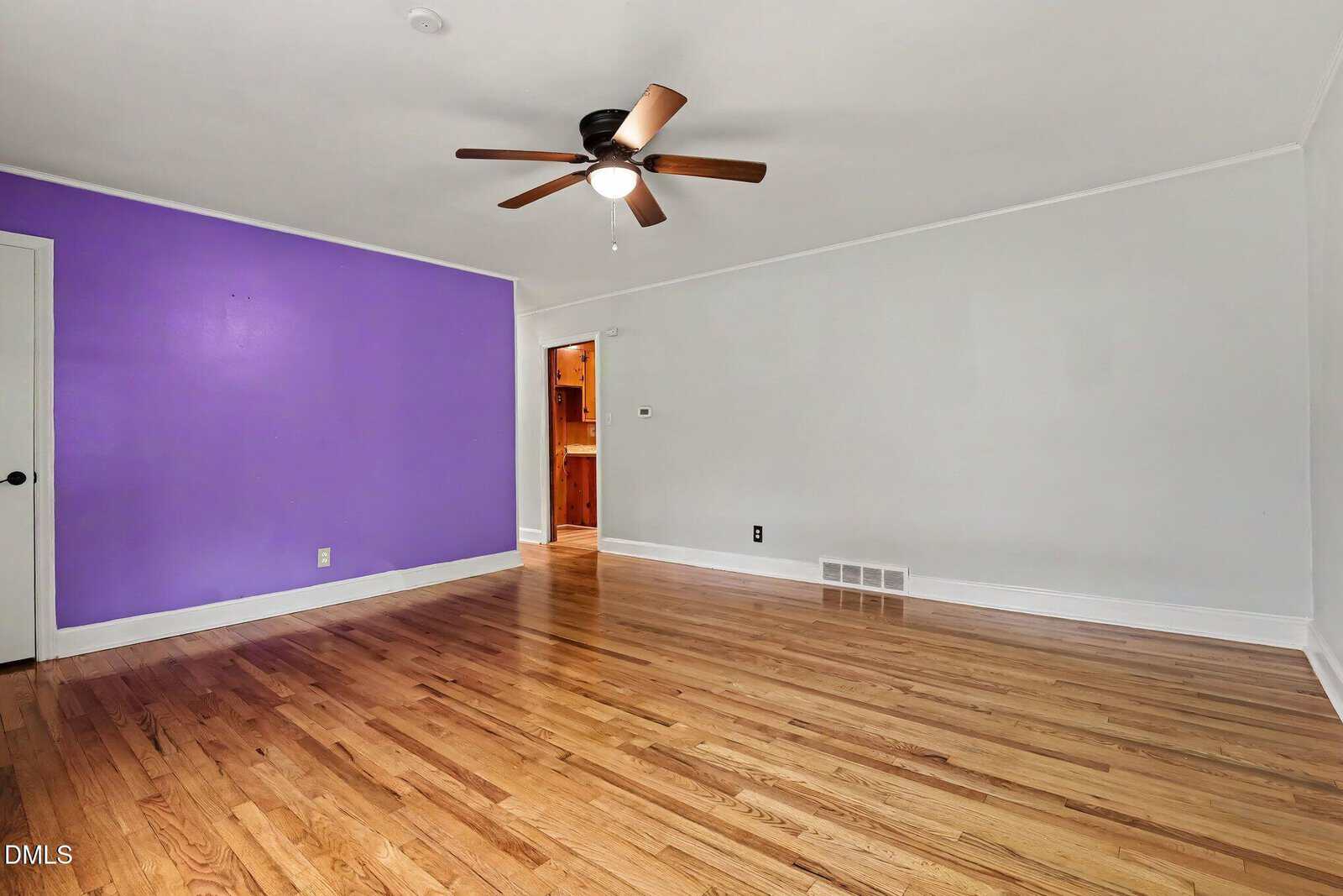 Empty room with light hardwood floors, white walls, one accent purple wall, a ceiling fan with light, a white door, and an open entrance revealing another room with wooden cabinets.
