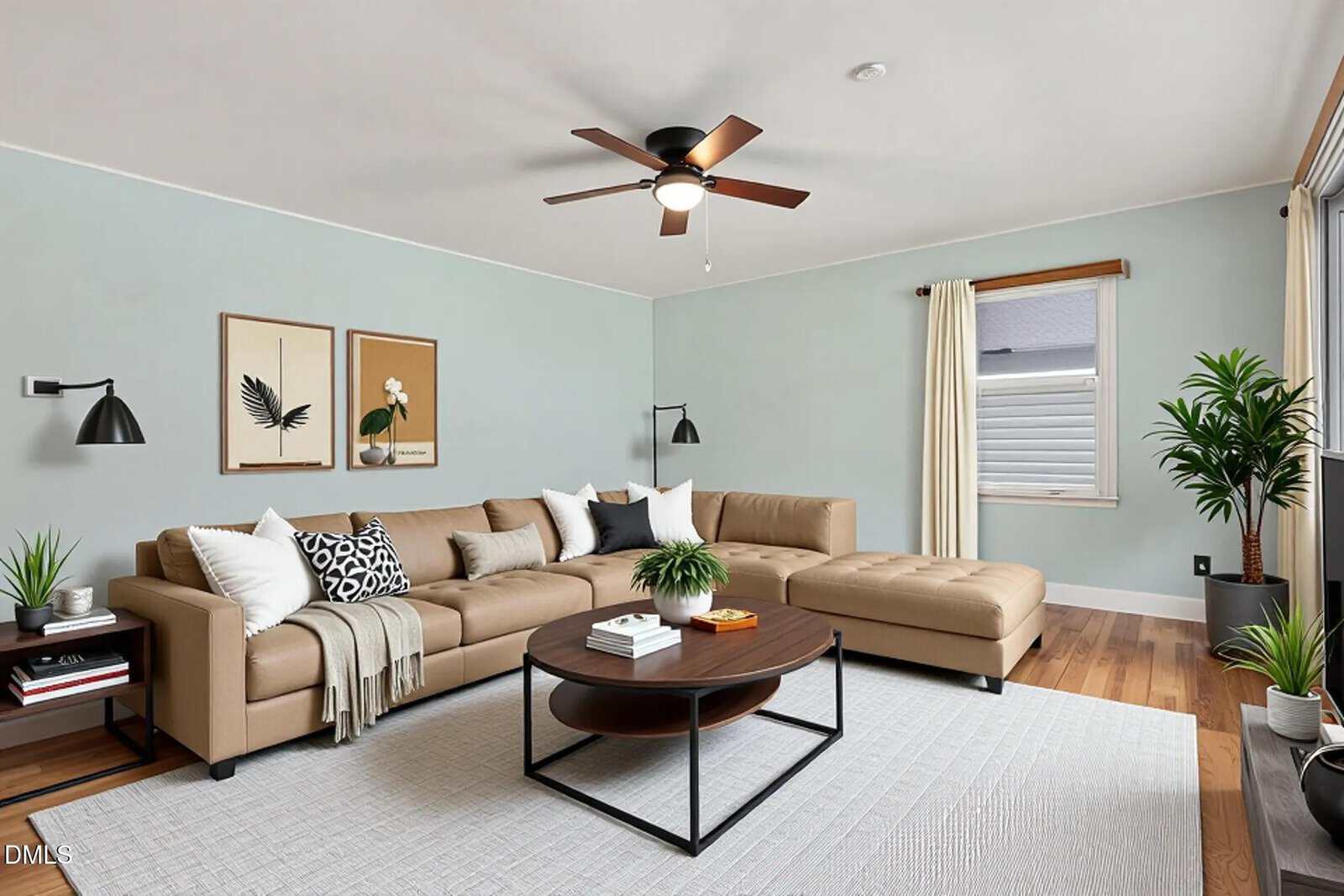 Modern living room with a beige sectional sofa, round coffee table, indoor plants, wall art, and a ceiling fan. Light blue walls and a large window with cream curtains brighten the space.