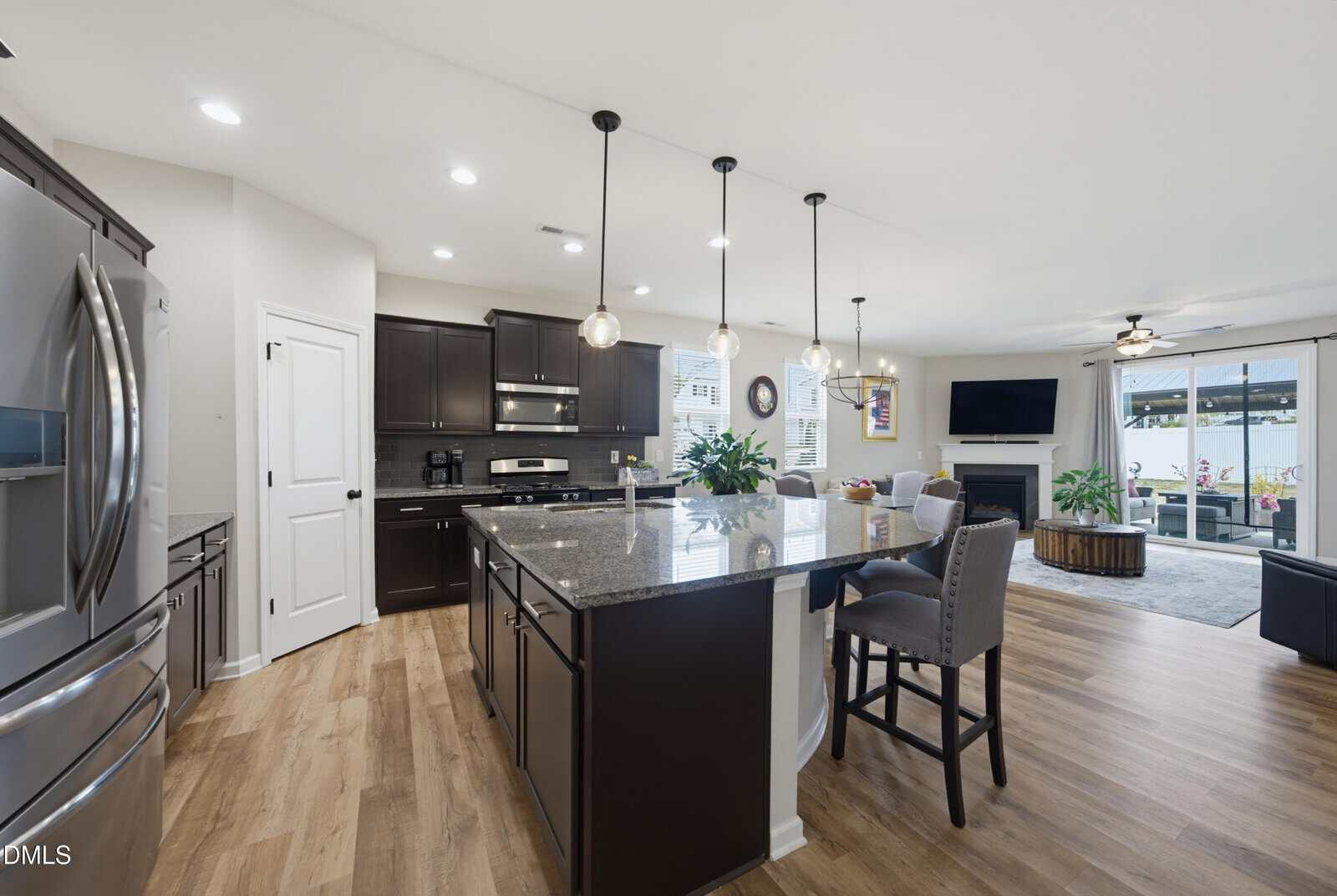 Modern open-concept kitchen and living area at 1036 Hazelmist Drive in Youngsville, featuring dark cabinets, granite island, stainless steel appliances, and wood flooring. Pendant lights hang over the island; the living room offers a TV and large windows for plenty of natural light.