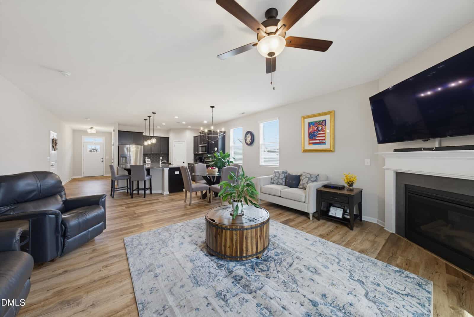 Modern open living room with wood floors, ceiling fan, dark kitchen cabinets, a round coffee table, blue-patterned rug, wall art, fireplace with mounted TV, and neutral-toned furniture. Bright and spacious atmosphere.