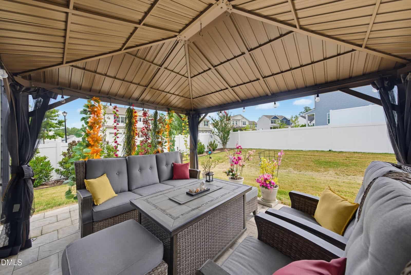 A cozy outdoor patio under a gazebo features wicker furniture with cushions, a table, potted flowers, and a grassy backyard enclosed by a white fence.