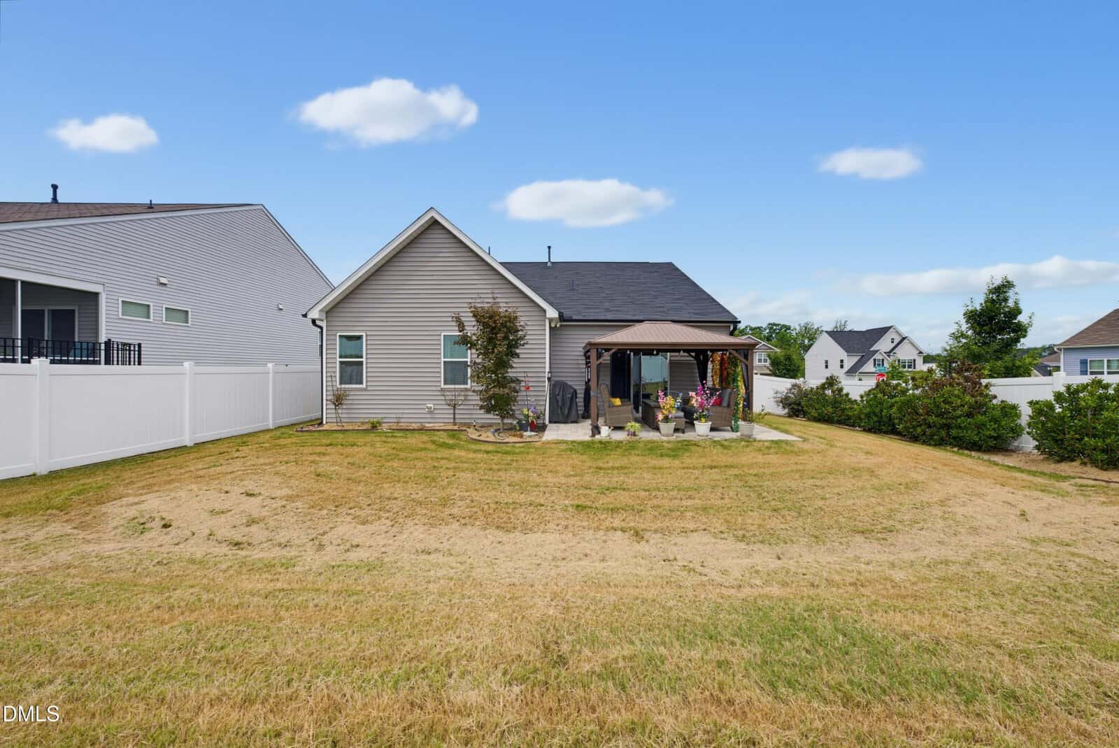 A backyard with a grassy lawn, a gray house, a small patio with a pergola, potted plants, and white fences on both sides under a blue sky with scattered clouds.