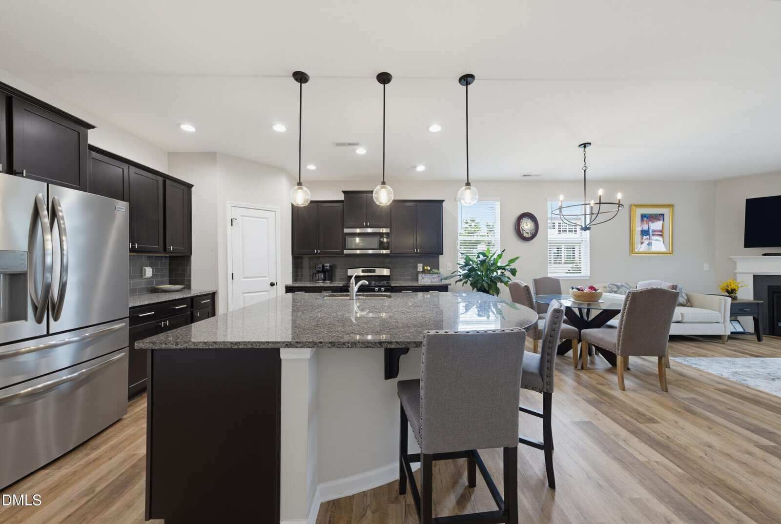 Modern kitchen with dark cabinets, stainless steel appliances, granite island, and three pendant lights. Open layout connects to dining area with a round table and chairs, and a living room with a fireplace and wall-mounted TV.