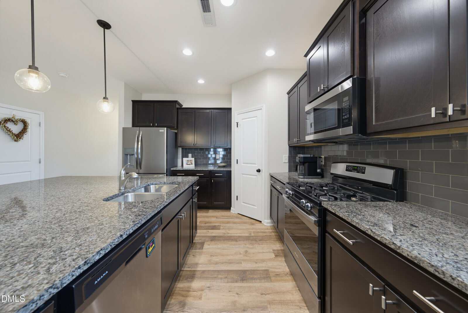 Modern kitchen with dark wood cabinets, stainless steel appliances, granite countertops, a center island with sink, pendant lights, light wood flooring, and a decorative heart on a white wall in the background.