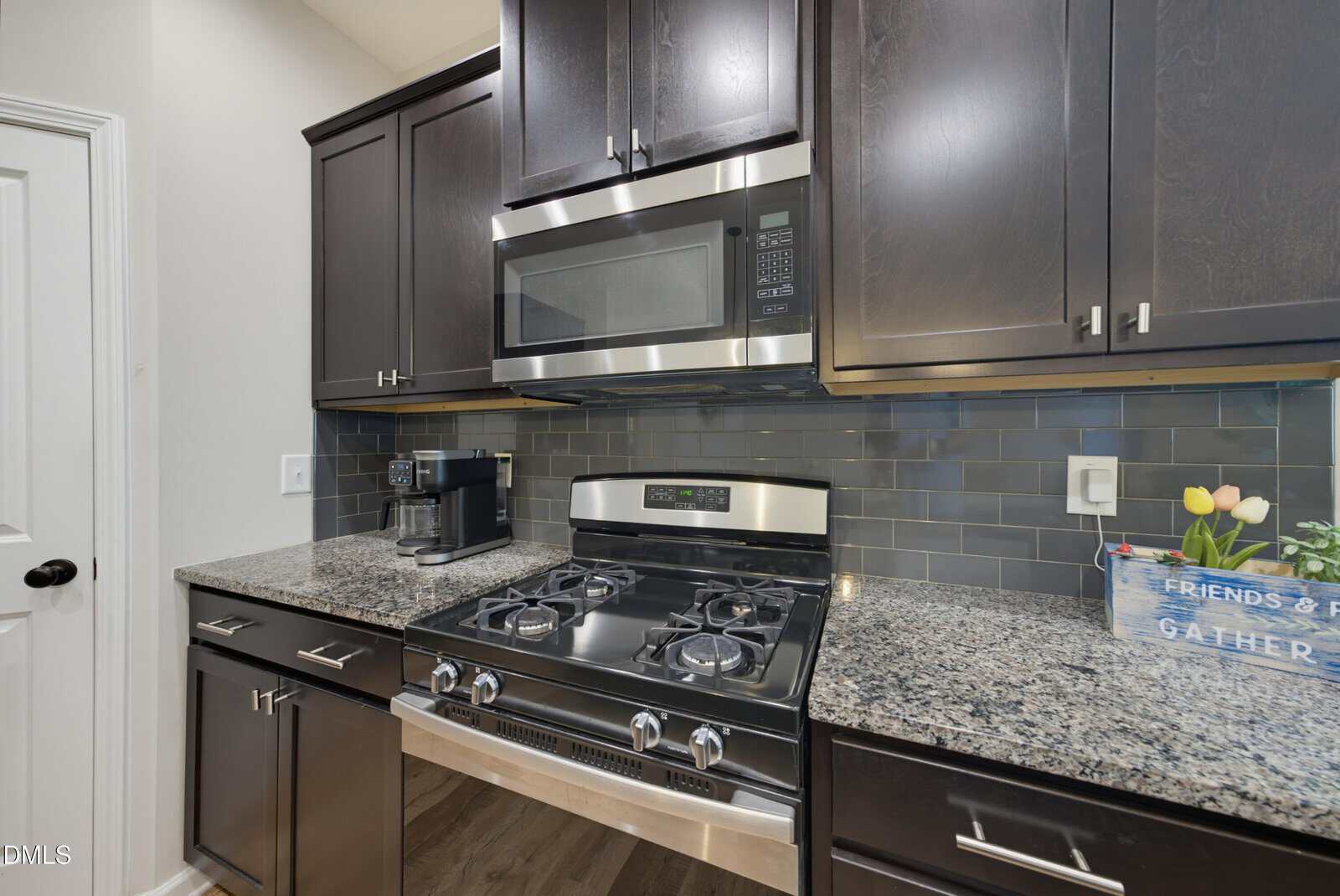A modern kitchen with dark cabinets, a stainless steel gas stove, a built-in microwave, granite countertops, a coffee maker, and a decorative tray with tulips on the right.