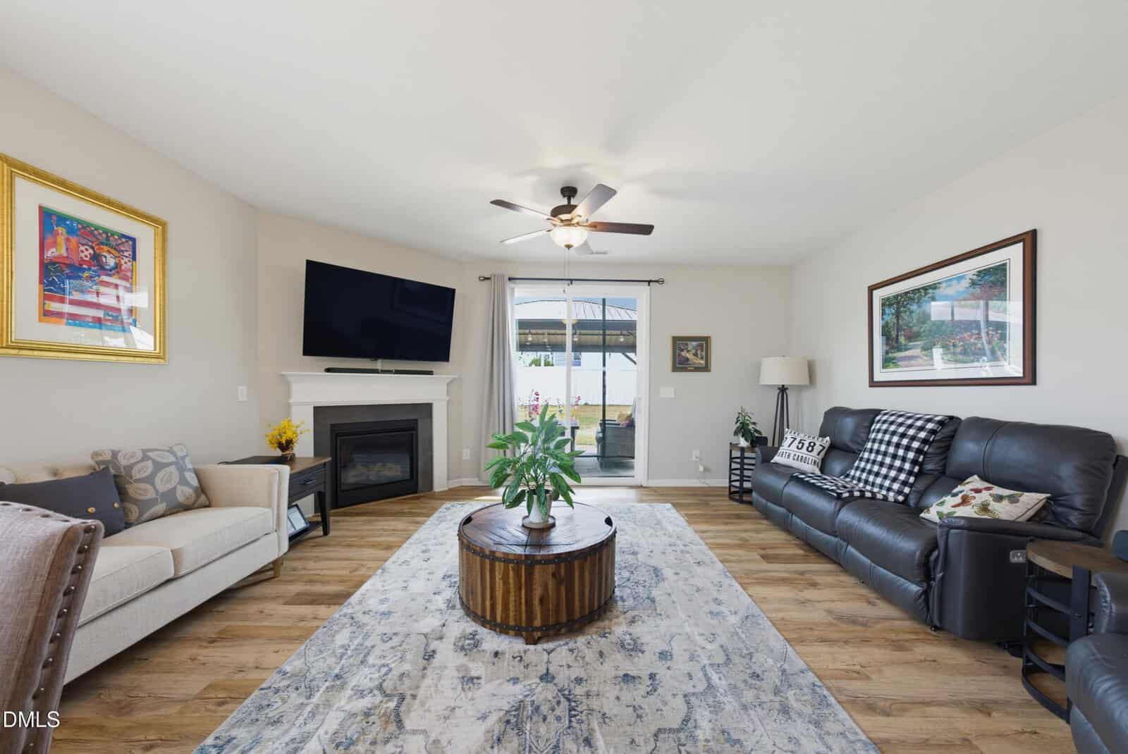Bright living room with a sofa, armchair, round coffee table, large TV above a fireplace, and a ceiling fan. Glass doors lead outside. Colorful art and houseplants add decor.