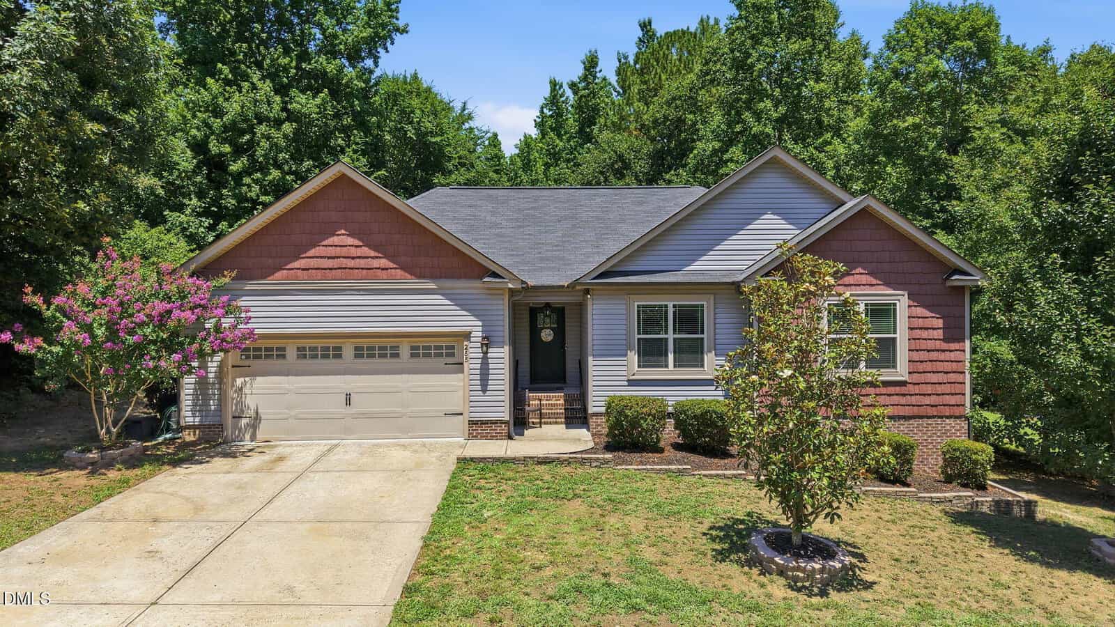 Charming single-story house at 265 N Ridge Drive, Louisburg, featuring beige and red siding, a two-car garage, a gray shingled roof, shrubs, a small tree in front, and surrounded by green trees under a clear sky.