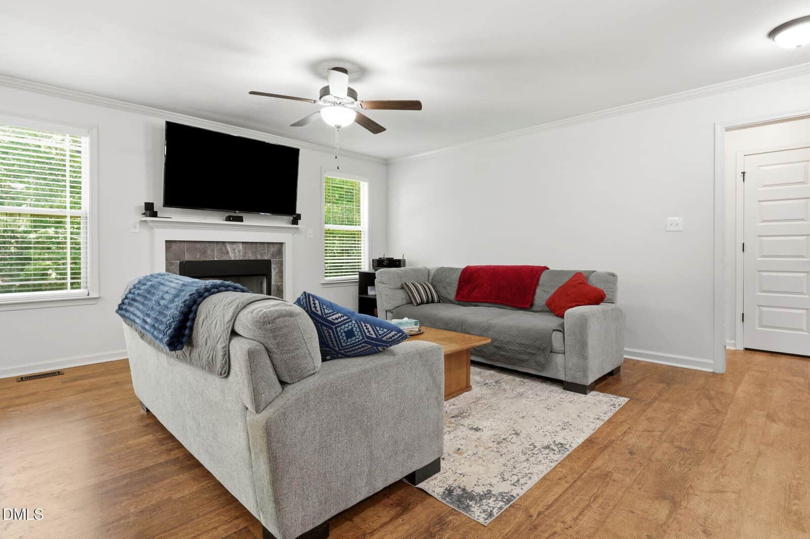 A bright living room with two gray sofas, a coffee table, a wall-mounted TV above a fireplace, and hardwood floors. Throw blankets and pillows add color to the neutral decor.