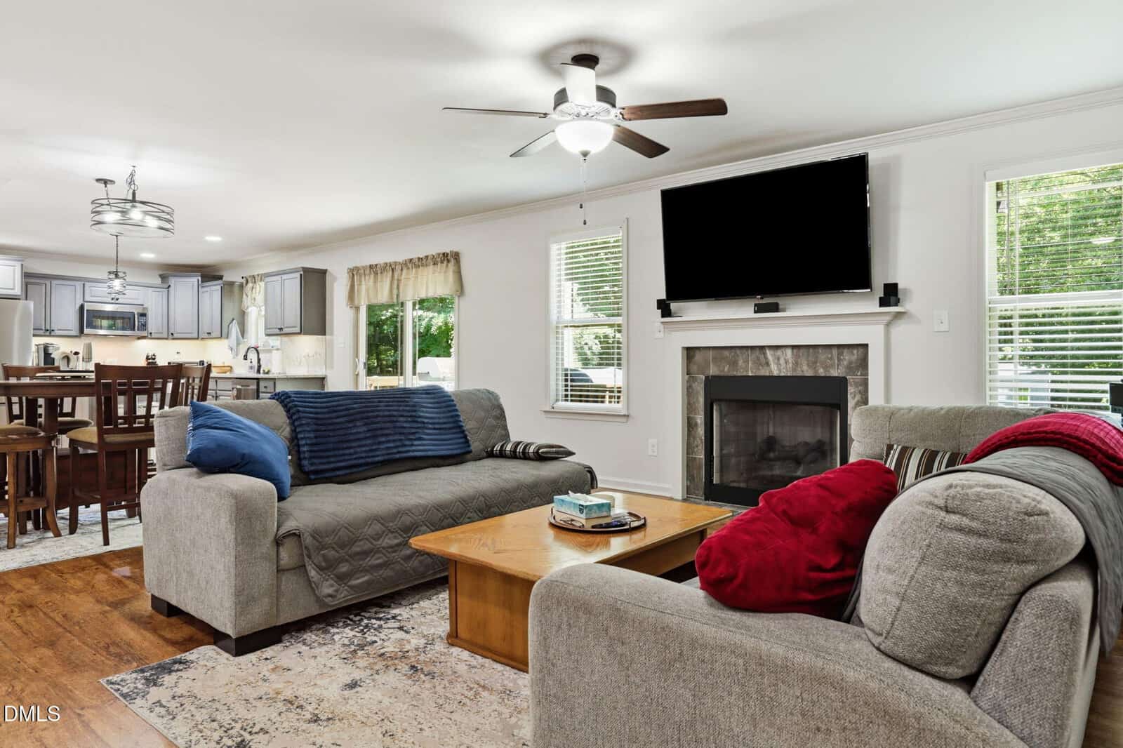 Modern open-concept living room with two gray sofas, colorful pillows, a wooden coffee table, TV above a fireplace, and a kitchen with barstools in the background. Large windows let in natural light.