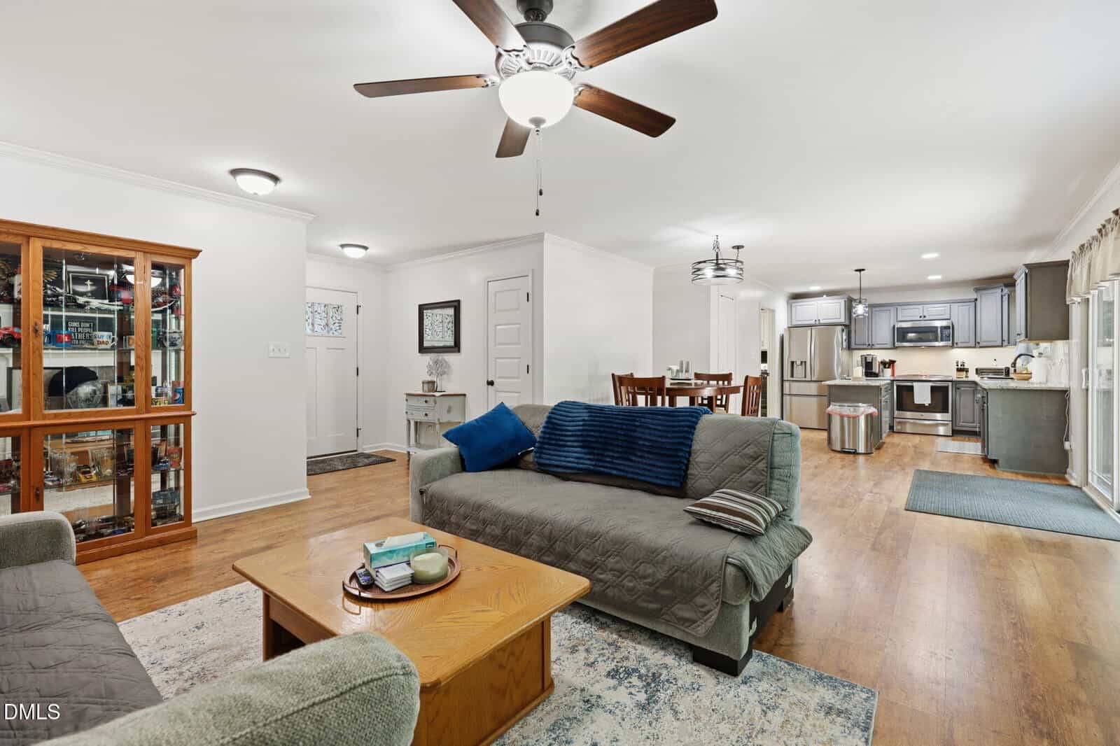 Open-concept living and dining area with wood floors, gray couches, a wooden coffee table, a dining table, and a modern kitchen in the background. Ceiling fans and light fixtures are visible throughout the space.