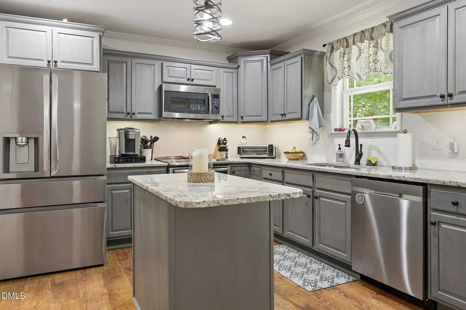Modern kitchen with gray cabinets, stainless steel appliances, marble countertops, an island with a candle centerpiece, a window with patterned curtains, and wooden floors. A rug is placed in front of the sink.